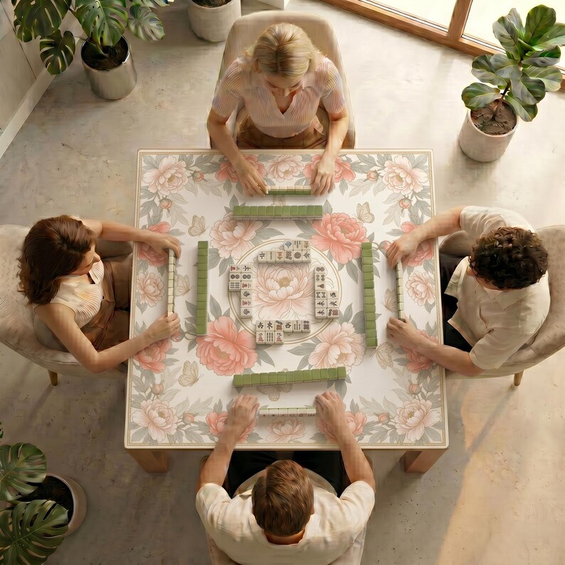 Overhead view of four people playing mahjong on a wooden table covered with the ivory peony mahjong mat in a bright room with potted plants.