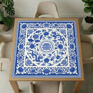 Blue and white dragon lotus mahjong mat covering a square wooden dining table, surrounded by neutral chairs and green indoor plants.