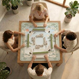 Overhead view of four people playing mahjong on a table covered with the cranes and pine clouds mahjong mat in a bright room with potted plants.