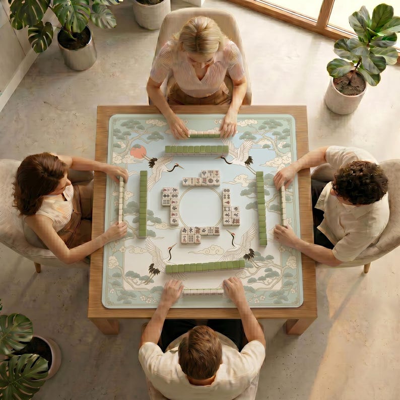 Overhead view of four people playing mahjong on a table covered with the cranes and pine clouds mahjong mat in a bright room with potted plants.