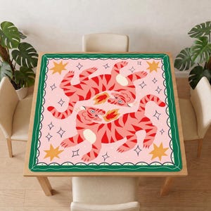 May include: A square tablecloth featuring a light pink background with two tigers in shades of red and pink. Each tiger has a white belly and is breathing a small orange flame. The design includes yellow stars and white four-pointed stars.