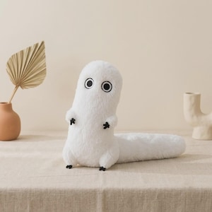 May include: A white plush toy with a long, cylindrical body and a rounded head. It features two large, circular black-and-white eyes, small black arms, and tiny black feet. The texture of the plush is soft and fuzzy, giving it a distinct appearance.