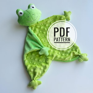 May include: A light green plush frog security blanket featuring a soft, textured minky fabric with raised dots. The frog head has embroidered eyes and a stitched mouth. A circular white graphic overlay displays the text PDF PATTERN.