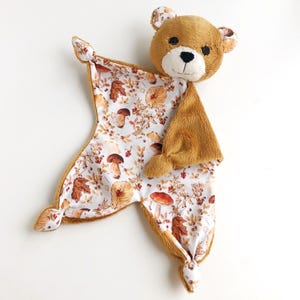 Könnte beinhalten: Eine Kuscheldecke mit einem braunen Teddybärenkopf aus weichem, flauschigem Material. Der Körper der Decke ist weiß und mit einem detaillierten herbstlichen Muster aus braunen Pilzen, getrockneten Blättern und kleinen roten Beeren bedruckt.