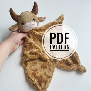 Könnte beinhalten: Eine kuschelige, braune und beige Baby-Sicherheitsdecke mit einem Kuhkopf. Die Decke hat ein Sternmuster und den Text "PDF PATTERN" in einem weißen Kreis. Die Kuh hat braune Hörner und Ohren mit einer weißen Schnauze.