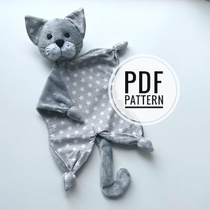 May include: A gray plush cat security blanket featuring a soft fabric body with a white star pattern, knotted corners, and a long tail. The head has embroidered black eyes, a black nose, and whiskers. A circular overlay displays the text PDF PATTERN.