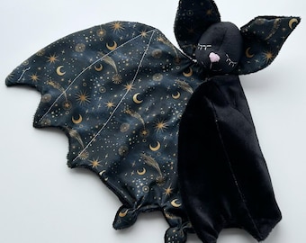 May include: A plush bat toy featuring a black head with a pink nose and embroidered eyes. One wing is made of black fabric with a gold celestial print of stars, moons, and comets, while the other wing is solid black plush fabric.