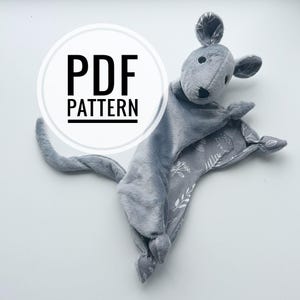 May include: A grey plush mouse security blanket with a soft texture, featuring a mouse head, a long tail, and knotted corners. The underside of the blanket and the inner ears display a white botanical and hedgehog print. Text reads PDF PATTERN.