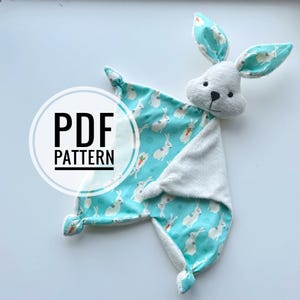 Puede incluir: Un arrullo para bebé con forma de conejito, con cabeza blanca y orejas con estampado de conejitos azules y blancos. El arrullo tiene un fondo turquesa con conejitos y zanahorias blancos. El texto "PDF PATTERN" está en un círculo blanco.