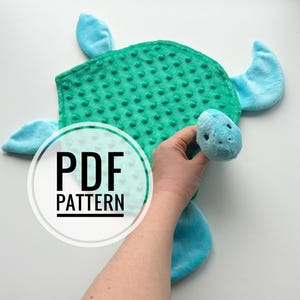 Peut inclure: Une peluche en forme de tortue avec une carapace en tissu vert texturé à pois et des nageoires ainsi qu'une tête en tissu bleu clair lisse. La tête présente de petits yeux brodés noirs. Un logo blanc circulaire avec le texte PDF PATTERN est visible.