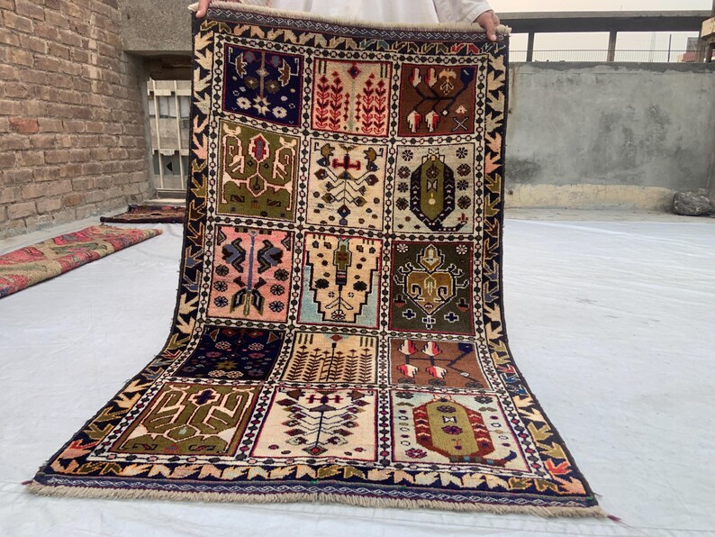 Pu&ograve; includere: Un tappeto rettangolare caratterizzato da una griglia di dodici pannelli distinti, ognuno dei quali mostra motivi geometrici e floreali unici in tonalit&agrave; blu navy, verde oliva, rosa, marrone e crema. Il tappeto &egrave; rifinito con frange alle estremit&agrave;.