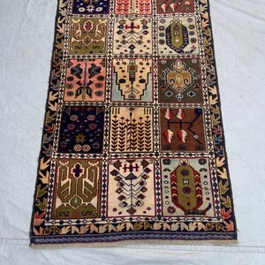 Pu&ograve; includere: Un tappeto rettangolare caratterizzato da una griglia di dodici pannelli distinti, ognuno dei quali mostra motivi geometrici e floreali unici nei toni del blu navy, verde oliva, terracotta, rosa e crema, racchiusi da un bordo scuro decorato.