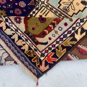 Pu&ograve; includere: Un tappeto piegato con un complesso motivo geometrico e floreale nei toni del blu navy, verde oliva, crema, marrone e rosso. Il tappeto presenta una consistenza a pelo spesso, un bordo decorativo con motivi a foglia e frange chiare.