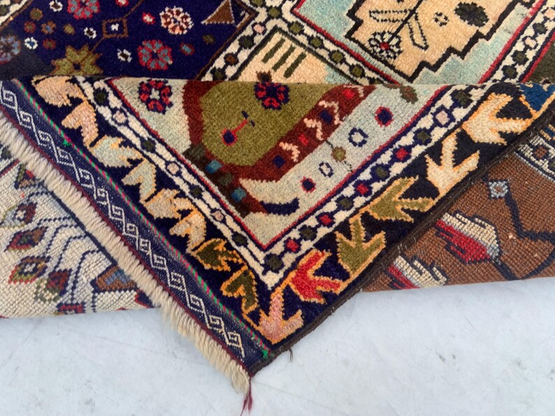 Pu&ograve; includere: Un tappeto piegato con un complesso motivo geometrico e floreale nei toni del blu navy, verde oliva, crema, marrone e rosso. Il tappeto presenta una consistenza a pelo spesso, un bordo decorativo con motivi a foglia e frange chiare.