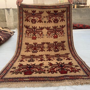 May include: A rectangular tan rug featuring a repeating pattern of dark red and blue floral motifs arranged in symmetrical rows. The rug has a decorative border with geometric shapes and includes fringe detailing along the top and bottom edges.