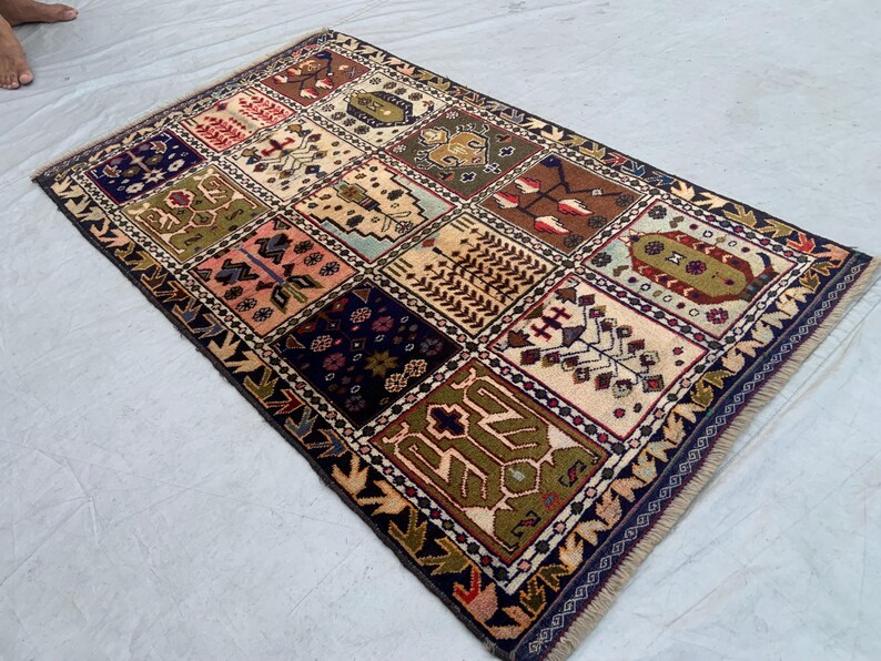Pu&ograve; includere: Un tappeto rettangolare caratterizzato da una griglia di dodici pannelli distinti, ognuno con motivi geometrici e floreali unici nei toni del blu navy, verde oliva, crema, marrone e rosa, rifinito con un bordo blu scuro e frange.