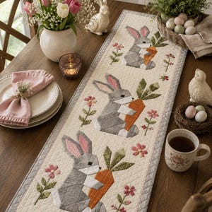 May include: A rectangular quilted table runner featuring a repeating pattern of three gray rabbits, each holding an orange carrot, interspersed with small pink and green floral motifs on a cream background with a gray border.