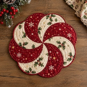 May include: A circular quilted fabric mat featuring a swirling pinwheel pattern. The design alternates between deep red sections with white snowflakes and cream-colored sections decorated with green holly leaves and red berries, finished with stitching.