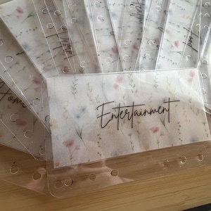 Op de afbeelding: Een set doorzichtige, rechthoekige plastic enveloppen voor een ringband, voorzien van een delicaat bloemenpatroon in zachte roze, groene en gele tinten. Elke envelop heeft voorgestanste gaten en zwarte, cursieve tekstlabels.