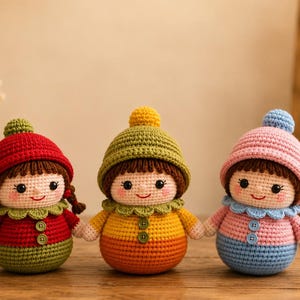 Puede incluir: Tres muñecas de ganchillo hechas a mano con cuerpos redondos, pelo castaño y gorros coloridos. La muñeca de la izquierda viste de rojo y verde, la del centro de oliva y naranja, y la de la derecha de rosa y azul. Tienen dos botones frontales.
