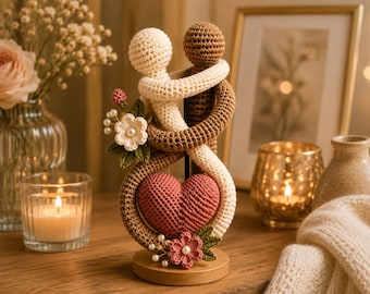 Op de afbeelding: Een gehaakt beeldje met twee figuren, één crème en één bruin, die elkaar omhelzen. Een groot roze hart is tussen hen geplaatst. De figuren zijn versierd met kleine gehaakte bloemen, groene blaadjes en witte parelachtige kralen, gemonteerd op een ronde houten voet.