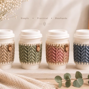 Op de afbeelding: Vier witte wegwerp koffiebekers met gestructureerde gehaakte hoezen in beige, saliegroen, oudroze en leiblauw. Elke hoes heeft een crèmekleurige rand, een klein bruin lipje in lederlook en een houten knoop. Tekst: Simple • Practical • Handmade.