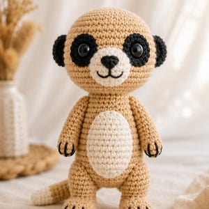 May include: A crocheted meerkat toy featuring a tan body, a cream-coloured belly, and black patches around its large dark eyes. The figure has small black ears, a black stitched nose and mouth, and black claw details on its hands and feet.
