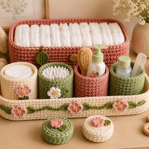 May include: A set of crochet storage organizers including a large rectangular basket, a long tray, four cylindrical containers, and two small round lids. The items feature cream, sage green, and dusty rose yarn with floral and leaf appliques.