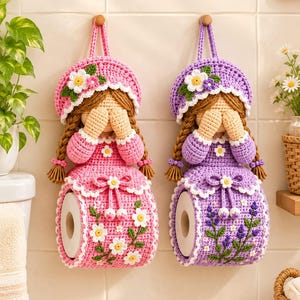 May include: Two handmade crochet toilet paper holders shaped like dolls with braided hair, one in pink with daisy accents and one in purple with lavender embroidery. Each doll wears a matching bonnet and covers its eyes with its hands.