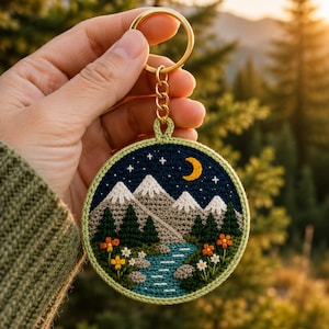 Op de afbeelding: Een ronde gehaakte sleutelhanger met een landschapstafereel met besneeuwde bergen, een donkerblauwe nachtelijke hemel met een gele maansikkel en witte sterren, een stromende rivier, groene dennenbomen en kleine oranje en witte bloemen.