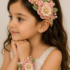 May include: A crochet headband and matching wrist cuff set featuring layered pink and cream flowers, green crocheted leaves, and small white pearl accents. The floral embellishments are attached to a delicate, open-work cream crochet band.