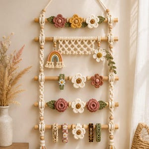 May include: A wall-hanging organizer featuring five horizontal wooden dowels suspended by macrame cord. The piece is decorated with crocheted flowers, a small rainbow charm, and various hair clips attached to the dowels, finished with tassels.
