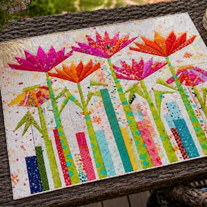 May include: A rectangular fabric wall hanging featuring a quilted floral design with bright pink and orange flowers on long green stems. The background is white with colorful paint splatters, and the base consists of vertical fabric strips in blue, green, pink, and yellow patterns.