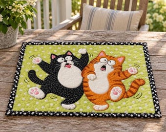 May include: A rectangular quilted fabric mat featuring a light green background with white polka dots. Two cartoon cats, one black and one orange tabby, are depicted in a startled pose with wide eyes and open mouths, framed by a black and white border.
