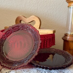 Puede incluir: Un juego de platos de vidrio rojo rubí con borde festoneado y un patrón decorativo de celosía en relieve alrededor del borde. El centro de cada plato muestra un diseño de rayos de sol y el vidrio tiene un color rojo profundo y translúcido.