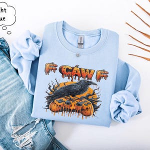May include: A light blue crewneck sweatshirt featuring a graphic print of a black crow perched on a melting orange pumpkin-shaped video game controller. The text "CAW" appears above the crow in a dripping orange font. A speech bubble says Light Blue.