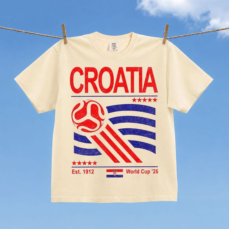 Puede incluir: Una camiseta de manga corta color crema con un estampado gr&aacute;fico en rojo y azul. El texto CROATIA aparece en la parte superior, junto a un bal&oacute;n de f&uacute;tbol, l&iacute;neas onduladas, cinco estrellas, Est. 1912, una bandera croata y World Cup '26.