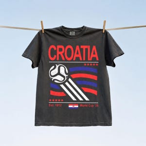 Puede incluir: Una camiseta de manga corta gris carb&oacute;n con el texto rojo CROATIA en la parte superior. Debajo, un gr&aacute;fico de bal&oacute;n de f&uacute;tbol blanco con l&iacute;neas de movimiento sobre rayas onduladas rojas y azules. Incluye cinco estrellas rojas, Est. 1912, una peque&ntilde;a bandera croata y World Cup '26.