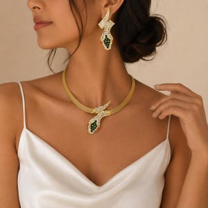 May include: A gold-toned snake-shaped necklace and matching earring set. The jewelry features a textured mesh band adorned with clear crystals, deep green stones, and small red accents. The snake head and tail designs are encrusted with stones.