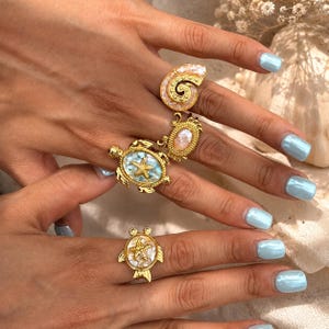 May include: Four gold-toned rings featuring nautical motifs. One ring is a spiral shell, one is an oval crab, one is a turtle with a starfish center, and one is a round crab with a starfish center. Each ring has a textured, iridescent inlay.