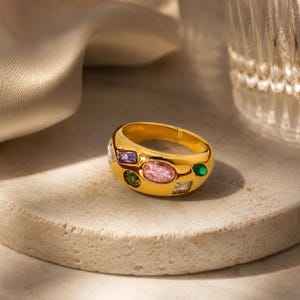 May include: A gold-toned ring featuring a thick, rounded band embedded with multiple multi-coloured gemstones. The stones include a large central oval pink gem, a rectangular purple gem, a small round green gem, and other clear stones.