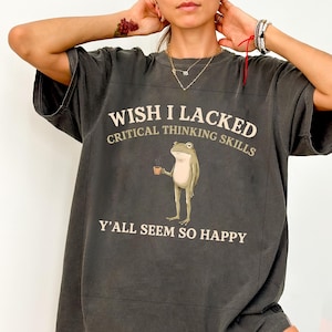 May include: A charcoal grey short-sleeve t-shirt featuring a graphic of a standing frog holding a small brown coffee mug. The text reads WISH I LACKED CRITICAL THINKING SKILLS Y'ALL SEEM SO HAPPY in cream-coloured lettering above and below the frog.