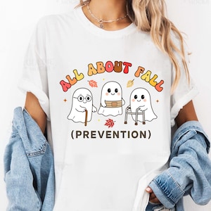 May include: A white t-shirt featuring a graphic of three cartoon ghosts with the text ALL ABOUT FALL (PREVENTION). One ghost wears glasses and holds a cane, one has a belt, and one uses a walker, surrounded by small colorful autumn leaves.