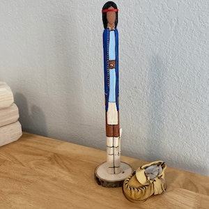 May include: A wooden figurine of a person wearing a blue dress, white skirt, and red headband. The figurine is standing on a wooden base and is next to a brown leather moccasin.