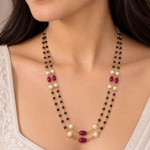 May include: A double-strand gold chain necklace featuring small black beads, white pearls, and oval-shaped magenta beads. The necklace is designed with a layered look, alternating the black beads with the white pearls and magenta accents throughout.