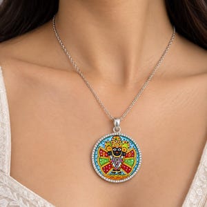 Könnte beinhalten: Eine runde silberne Anhängerkette mit einer farbenfrohen, detaillierten Illustration der Gottheit Jagannath. Der Anhänger ist von einem Rand aus kleinen, klaren Steinen umgeben und hängt an einer schlichten Silberkette vor weißem Stoff.