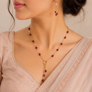 May include: A gold chain necklace featuring alternating small white pearls and oval-shaped red beads, culminating in a central circular gold ring with three dangling gold chains tipped with red beads, paired with matching drop earrings.