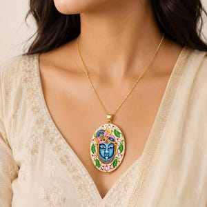 Könnte beinhalten: Eine oval geformte Anhängerkette mit einem handbemalten blauen Gottheitsgesicht und farbenfrohem Kopfschmuck, umgeben von einer dekorativen Bordüre aus rosa Lotusblüten und grünen Blättern auf cremefarbenem Grund, an einer dünnen goldfarbenen Kette.