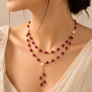 Puede incluir: Un collar de doble hebra con cuentas ovaladas de color rojo intenso y pequeñas perlas blancas unidas por una fina cadena dorada. El collar incluye un colgante central compuesto por varias cadenas doradas con pequeñas cuentas rojas.
