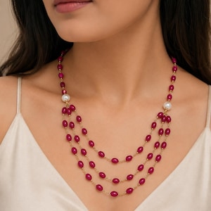 May include: A three-strand necklace featuring small, oval-shaped magenta beads strung on a thin gold-tone chain. The design includes two larger, round white pearls positioned symmetrically on each side, connecting the three cascading strands.